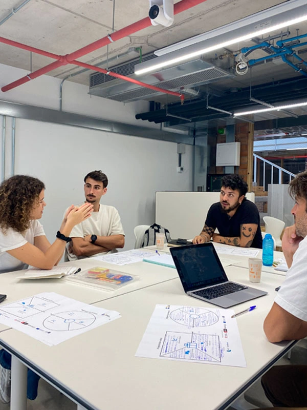 Persones realitzant una sessió de treball col·laborativa al voltant d’una taula, amb documents de treball i un ordinador portàtil.