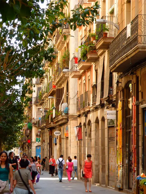 Persones caminant per un carrer urbà amb edificis antics, balcons i comerços a peu de carrer.
