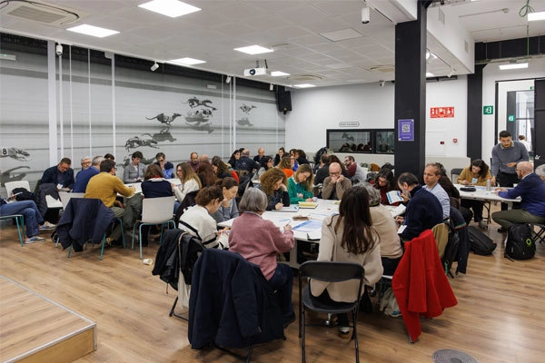 Persones treballant en grups al voltant de diverses taules durant una sessió participativa en un espai de formació.