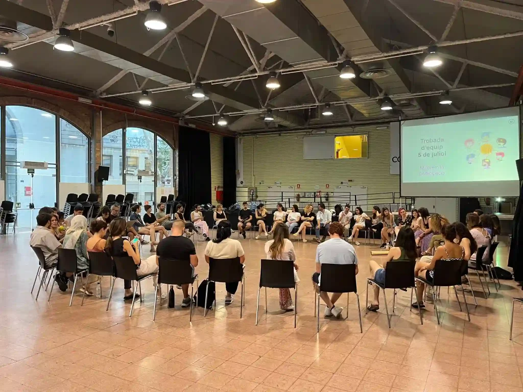 Grup nombrós de persones assegudes en rotllana durant una sessió grupal en un espai ampli, amb una presentació projectada al fons.
