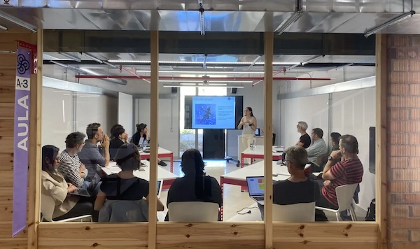 Un grup de persones adultes dins una aula en rotllana escoltant l’explicació de la professora.